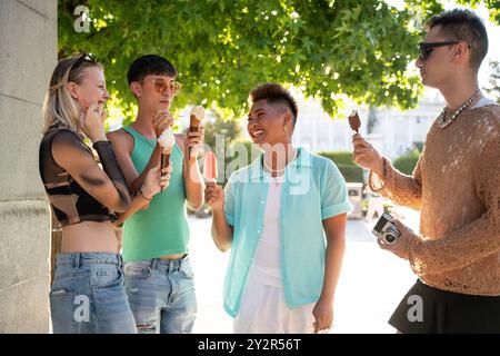 Eine Gruppe von vier LGBTQIA+ Freunden steht unter einem Baum, plaudert und lacht, während sie Eiskegel und Eis genießen. Ihr Glück und ihre Kameradschaft Stockfoto