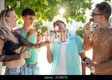 Eine Gruppe von vier LGBTQIA+ Freunden steht unter einem Baum, plaudert und lacht, während sie Eiskegel und Eis genießen. Ihr Glück und ihre Kameradschaft Stockfoto