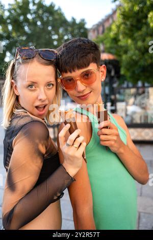 Zwei LGBTQIA+ Freunde genießen gemeinsam Eiskegel in einer lebhaften Stadtkulisse. Sie lächeln, tragen eine stilvolle Sonnenbrille und drücken Freude und aus Stockfoto