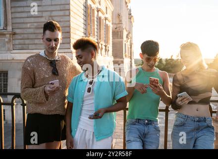 Vier LGBTQIA+-Freunde genießen einen fröhlichen Moment zusammen, während sie auf einer sonnigen Stadtterrasse auf ihren Telefonen surfen, was Vielfalt und Freundschaft widerspiegelt. Stockfoto