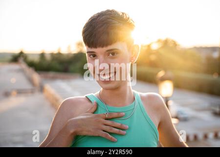 Schwuler junger Mann mit einem stilvollen Haarschnitt, der während eines wunderschönen Sonnenuntergangs in einem lässigen Tanktop warm lächelt Stockfoto