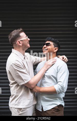 Fröhliches, multiethnisches schwules Paar, das sich von Herzen lacht, eng mit Armen, die sich umarmen, vor einem auffälligen schwarzen strukturierten B steht Stockfoto