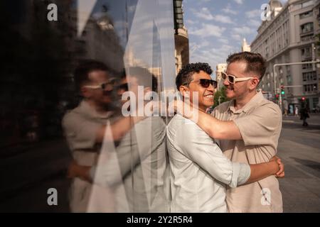 Ein liebevolles, multiethnisches schwules Paar umarmt sich herzlich auf einer belebten Straße, ihr Lachen und ihre Freude in der Reflexion eines Glasgewinns Stockfoto