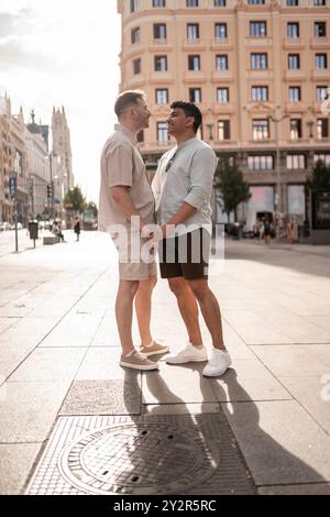 Ein liebevolles, multiethnisches schwules Paar, das Hände hält und ein Lächeln in einer hell beleuchteten urbanen Umgebung mit klassischer Architektur im Hintergrund teilt. Stockfoto