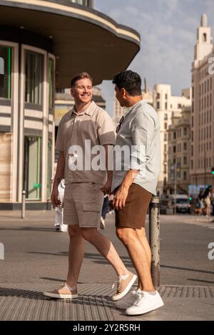 Ein multiethnisches schwules Paar teilt einen freudigen Moment in einer sonnigen Stadtstraße und führt in liebevolle Gespräche, umgeben von urbaner Architektur. Stockfoto