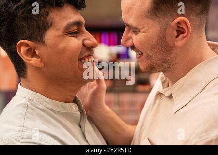 Ein zärtliches und Nahaufnahme-Bild, das ein fröhliches multiethnisches schwules Paar in einem Moment der Zuneigung einfängt, warm lächelt und sich gegenseitig die Gesichter berührt. Stockfoto