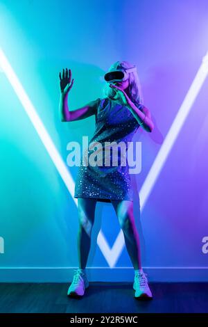 Eine Frau in einem glitzernden Kleid und Turnschuhen verwendet ein VR-Headset gegen leuchtende blaue und violette Lichter und simuliert so futuristische Metaversen. Stockfoto