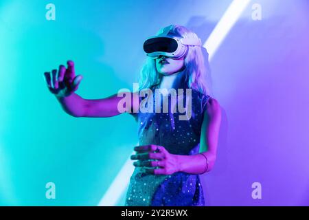 Eine junge Frau in einem Kleid mit Punktmuster verwendet ein VR-Headset unter leuchtenden Neonlichtern und symbolisiert damit, dass sie in das Metaverse eintaucht. Ihr Ausdruck Stockfoto