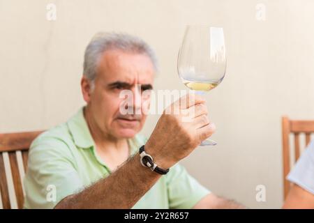 Ein reifer Mann in einem hellgrünen Hemd inspiziert sorgfältig ein Glas Weißwein und teilt vielleicht einen Moment mit jemandem, der nicht im Rahmen ist Stockfoto