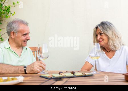 Ein reifes Paar sitzt an einem Holztisch im Freien, lächelt und genießt Weißweingläser, umgeben von einer warmen, ruhigen Umgebung Stockfoto