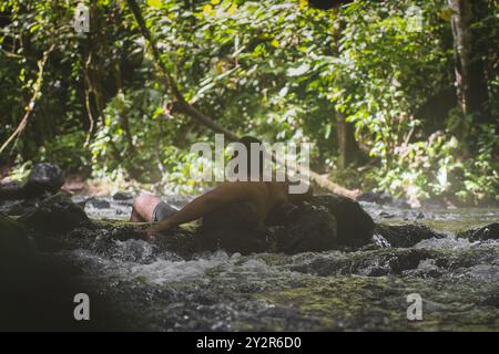 Ein Mann wird gezeigt, wie er sich in einem klaren, fließenden Fluss inmitten einer üppigen Vegetation in Costa Rica entspannt, was einen ruhigen Rückzug in die Natur veranschaulicht Stockfoto