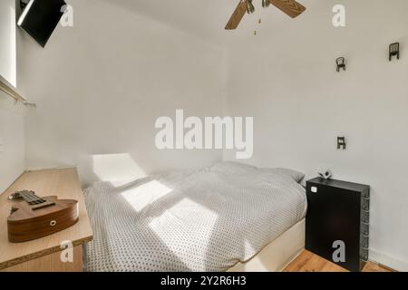 Ein gut beleuchtetes, gemütliches kleines Schlafzimmer mit einem Bett mit Punktlaken, einer Gitarre, einem Deckenventilator und minimalistischer Wandkunst. Stockfoto