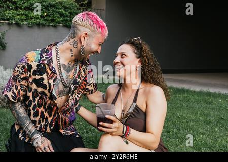 Ein trendiges junges Paar teilt einen freudigen Moment bei einem Drink in einer grasbewachsenen Umgebung im Freien. Der Mann, mit rosa Haaren und Tattoos geschmückt, lächelt aufrichtig an Stockfoto