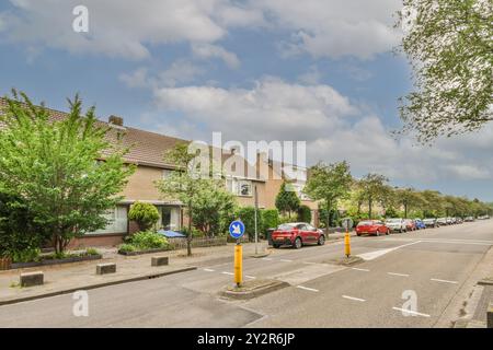 Eine friedliche Vorstadtstraße gesäumt von Reihenhäusern, üppigem Grün und einem klaren blauen Himmel mit geparkten Autos und Straßenschildern. Stockfoto