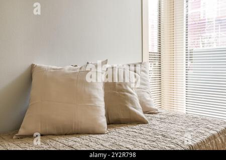 Eine ruhige Schlafzimmerecke mit drei Leinenkissen auf einer strukturierten Decke, beleuchtet von sanftem Sonnenlicht, das durch venezianische Jalousien gefiltert wird. Stockfoto