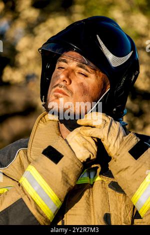 Ein Nahbild eines Feuerwehrmannes in Schutzausrüstung, der seinen Helm unter goldenem Sonnenlicht einstellt, mit verschwommenen Bäumen im Hintergrund Stockfoto