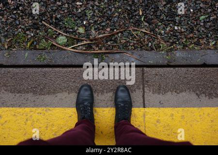 Suchen Sie nach dem Boden an einem antiken Bahnhof an einem regnerischen Tag. Schwarze Lederstiefel über der gelben Linie der Plattform. Stockfoto