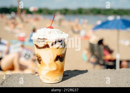 Ein köstlicher Eisbecher mit Schlagsahne und einer Kirsche an einem sonnigen Strand, an dem sich die Leute im Hintergrund entspannen Stockfoto