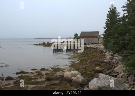 Ein rustikales Bootshaus in der Nähe von Peggy's Cove, Nova Scotia, mit einem felsigen Ufer, ruhigem Wasser und einem bewölkten Himmel. Stockfoto