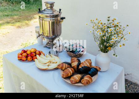 Ein wunderschönes Frühstück im Freien wird mit einem traditionellen Samovar, Croissants und Gebäck auf einem Tisch serviert, zusammen mit frischem Obst und einer Vase Stockfoto