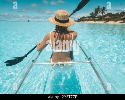 Eine Frau, die in einem klaren, transparenten Kajak an einem tropischen Strand mit türkisfarbenem Wasser und Palmen Kajak fährt Stockfoto
