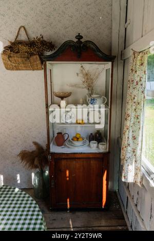 Ein Holzschrank steht in einer sonnigen Landküche, in der eine Auswahl an Vintage-Gerichten, dekorativen Stücken und frischen Zitronen präsentiert wird, was eine warme, inviente Atmosphäre schafft Stockfoto