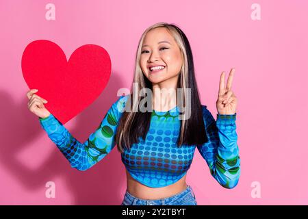 Foto von fröhlicher hübscher Dame gekleidetes blaues Hemd aufsteigendes rotes Herz, das V-Zeichen leeren Raum zeigt isolierten rosafarbenen Hintergrund Stockfoto