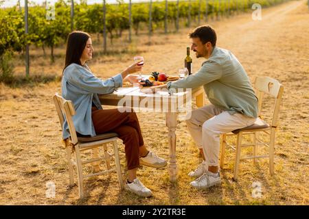 Ein paar entspannen sich an einem rustikalen Tisch in einem sonnigen Weinberg, genießen Wein und Gourmet-Snacks. Sie teilen Lachen und Freude, umgeben von üppigen Weinstöcken und dem Stockfoto