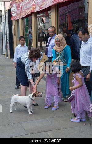 Eine weiße britische Frau und ihr Hund an der Leine werden gestreichelt, von einem jungen asiatischen Mädchen berührt, das bei ihrer Familie ist. Die Frau hält die Asiaten die Hand und hilft ihr, den Kopf des Hundes zu berühren. Brick Lane, East London, Whitechapel, London, England 30. Juni 2013 2010, UK HOMER SYKES Stockfoto