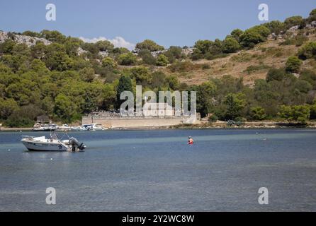 Eine ruhige Küstenszene mit einem Boot auf ruhigem Wasser mit einem historischen Gebäude und üppigen grünen Hügeln im Hintergrund. Stockfoto