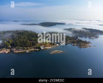 Morgennebel, der im Frühherbst auf einer Insel im schwedischen Archipel in Schweden über eine Insel rollt. Häuser, Bäume im Nebel. Blauer Himmel, helles Sonnenlicht Stockfoto
