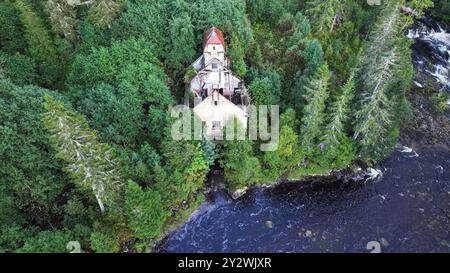 Ein Blick aus der Vogelperspektive auf eine alte Holzhütte in der Nähe eines neuen Damms und eines Flusses und eines Kraftwerks in einem grünen Wald Stockfoto