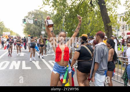 A woman in a red top and denim shorts raises her arms, holding a large bottle in one hand, as she joyfully participates in the West Indian Day Parade. Stockfoto