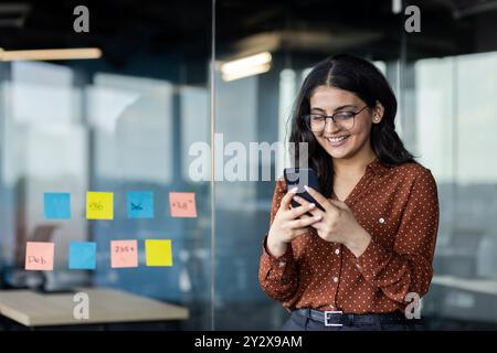 Junge Geschäftsfrau, die im Büro telefoniert, Firmenangestellte lächelnd SMS schreibt und soziale Netzwerke durchsucht, hispanische Frau zufrieden mit der Arbeit, die in der Nähe von Fenstern steht. Stockfoto
