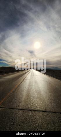 Eine lange, gerade Straße, die in die Ferne führt, unter einem dramatischen Himmel, in dem die Sonne hell scheint. Stockfoto