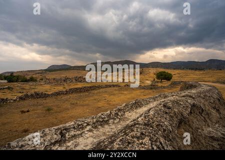 Hierapolis in Pamukkale: Ein einsamer Pfad schlängelt sich durch die trockene Landschaft und führt zu antiken Ruinen, die sich unter einem dramatischen, wolkenreichen Himmel befinden. Stockfoto