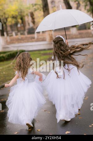 Eine Rückaufnahme von zwei Mädchen in langen weißen Festkleidern, die mit Regenschirm auf einer Straße entlang laufen Stockfoto