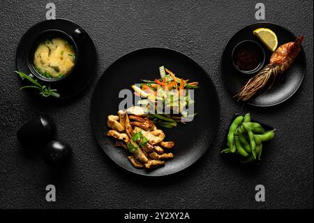 Ein wunderschön arrangiertes japanisches Menü mit Miso-Suppe, gegrillten Garnelen, Edamame und gebratenem Gemüse auf einem strukturierten schwarzen Hintergrund. Stockfoto