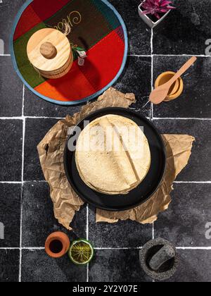 Blick von oben auf traditionelle mexikanische Tortillas auf einem schwarzen Teller mit verschiedenen Küchenartikeln auf einer gefliesten Oberfläche Stockfoto