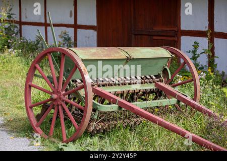 Eine altmodische grüne und rote Bohrmaschine auf Gras, mit einem rustikalen Holzgebäude im Hintergrund. Die Sämaschine verfügt über eine große Holzkeule Stockfoto