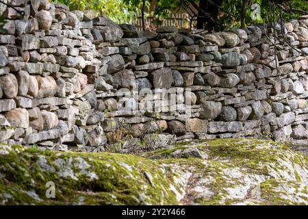 Eine rustikale Steinmauer aus verschiedenen Größen von Felsen, die eine natürliche, erdige Ästhetik zeigt. Die Mauer ist teilweise mit Moos bedeckt, umgeben von gre Stockfoto