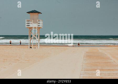 Leerer Strand mit weißer Rettungswache am Horizont am blauen mittelmeer. Holzhütte am morgendlichen Sandstrand. Holzweg führt zum Wasser. Stockfoto