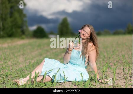 Eine junge Frau in einem hellblauen Kleid, die auf einem Feld sitzt, eine Blume hält, mit einem bewölkten Himmel im Hintergrund. Stockfoto
