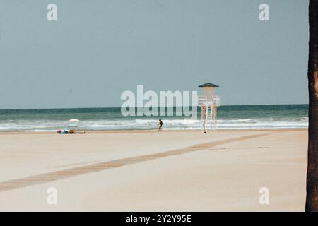 Leerer Strand mit weißer Rettungswache am Horizont am blauen mittelmeer. Holzhütte am morgendlichen Sandstrand. Holzweg führt zum Wasser. Stockfoto