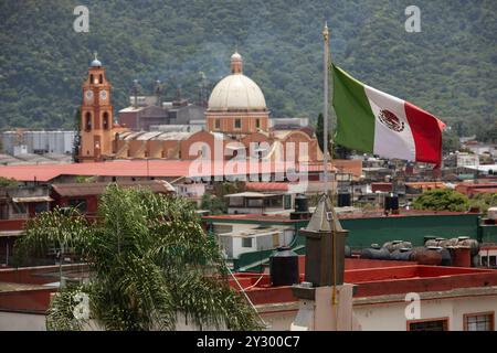 Orizaba, Veracruz, Mexiko - 14. Juli 2022: Nachmittagswind weht eine mexikanische Flagge und raucht von der Industrie neben einer Kirche in der Innenstadt von Orizaba. Stockfoto