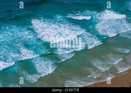 Aus der Vogelperspektive auf windgepeitschte Meereswellen, mehr weiß als blau, die zum Strand hin Rollen. Stockfoto