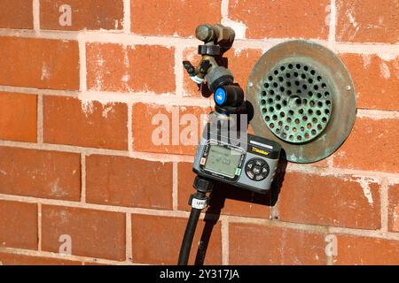 Ein Melnor hydrologischer Einzonen-Wasserzähler, der an einem Schlauch und einem Wasserhahn befestigt ist und an einer orangen Ziegelwand sitzt. Stockfoto