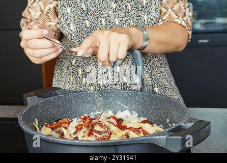 Eine Person fügt Gewürze mit einem Löffel in eine Pfanne mit sautйed-Zwiebeln und sonnengetrockneten Tomaten. Stockfoto