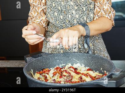 Eine Person fügt Gewürze mit einem Löffel in eine Pfanne mit sautierten Zwiebeln und sonnengetrockneten Tomaten. Stockfoto