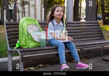 Glückliches Mädchen mit Rucksack, das auf der Parkbank sitzt und in Notizbuch schreibt Stockfoto